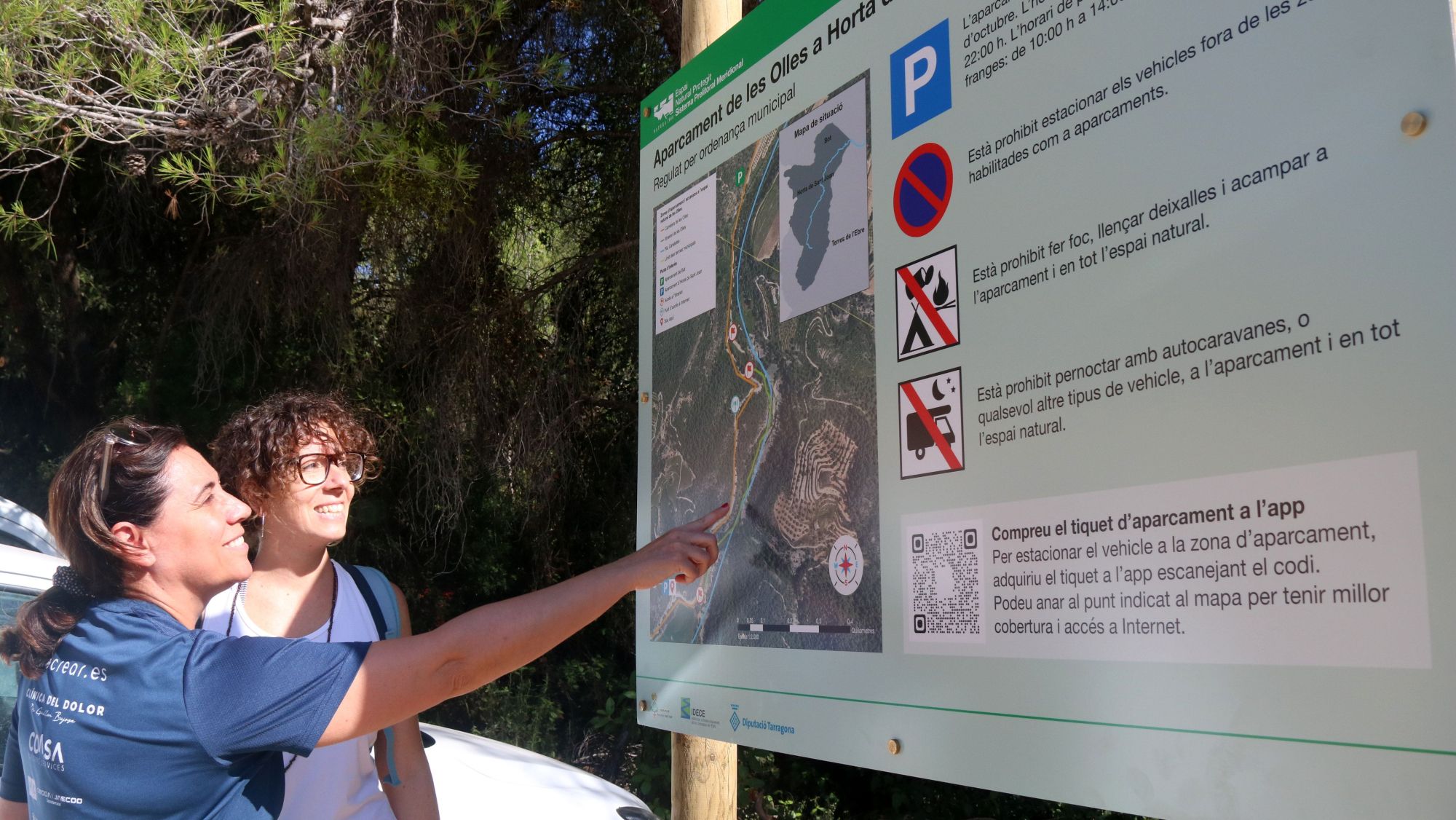 Dues usuàries revisen el cartell informatiu a l'aparcament regulat d'Horta de Sant Joan per accedir a l'espai de les Olles