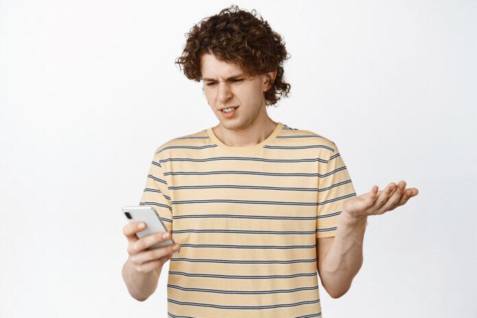 Image of curly guy staring confused at mobile phone, standing in t-shirt over white background