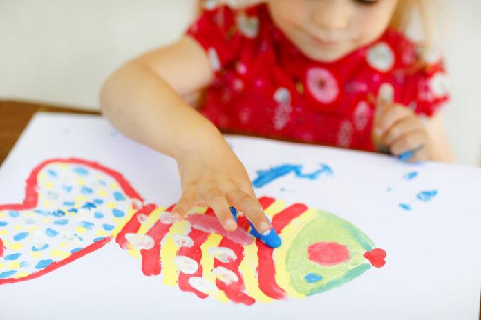 Little creative toddler girl painting with finger colors a fish. Active child having fun with drawing at home, in kindergaten or preschool. Education and distance learning for kids. Creaitve activity.